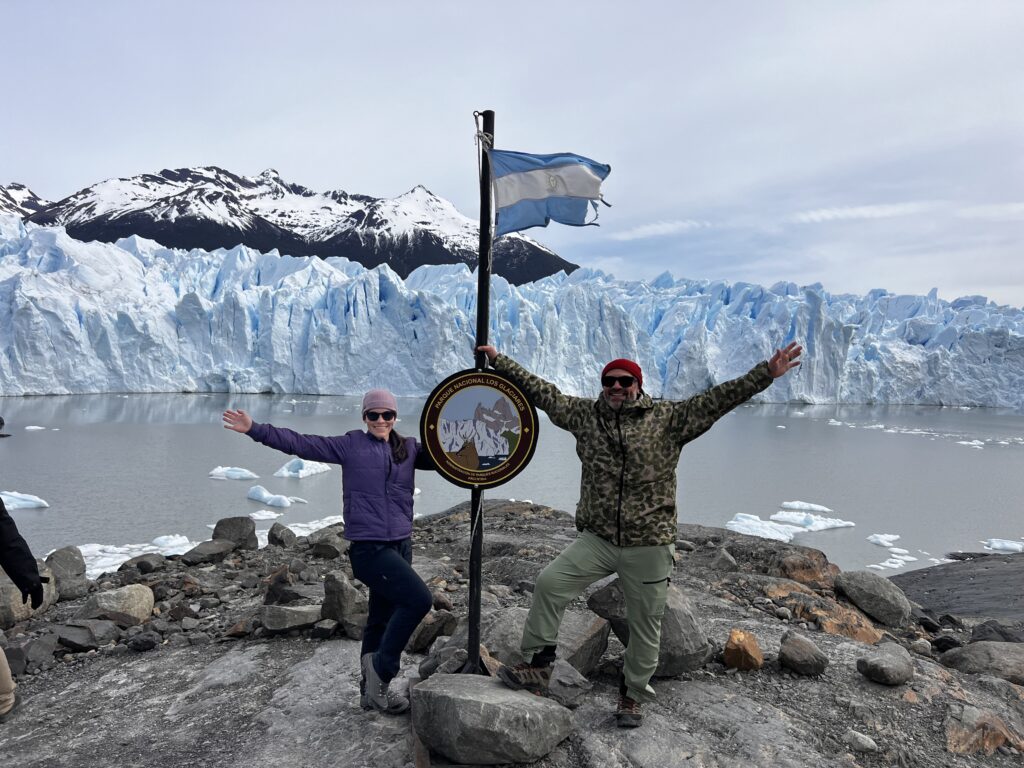 Two individuals pose in front of Perito Moreno Glacier, surrounded by jagged ice formations and snow-capped mountains. Argentine flag and "Parque Nacional Los Glaciares" sign add patriotic touch. Dress warmly for rocky terrain and floating ice chunks.